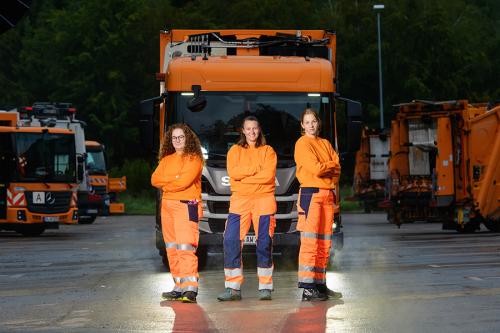 Drei Müllwerkerinnen vor mehreren Müllsammelfahrzeugen